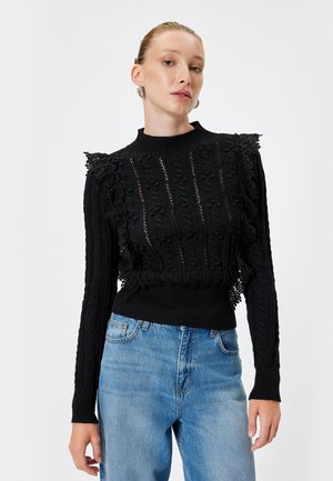 Schwarzer gestrickter Pullover mit langen Ärmeln, der strukturierte Muster und Spitzenakzente an den Schultern aufweist, kombiniert mit hellblauen Jeans.