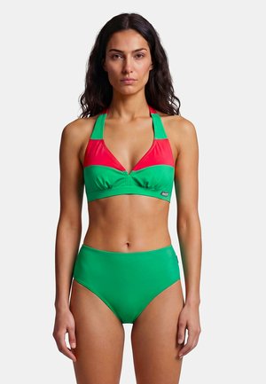 Frau mit langen dunklen Haaren, die ein grünes und rotes Neckholder-Bikinitop und grüne Bikinihosen mit hoher Taille trägt, steht vor weißem Hintergrund.