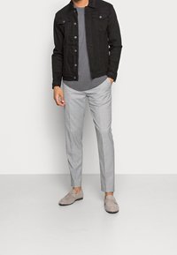 Uomo che indossa una giacca di jeans nera, una maglietta grigia, pantaloni eleganti grigio chiaro e mocassini in suede beige, in piedi contro uno sfondo semplice.