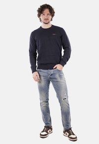 Maglione navy a girocollo con polsi a costine, abbinato a jeans chiari strappati e scarpe da ginnastica marroni con dettagli bianchi.