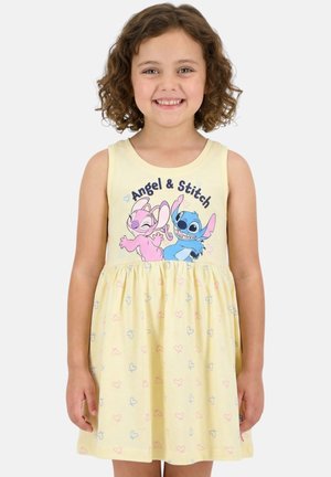 Fille souriante aux cheveux bouclés portant une robe jaune sans manches avec les personnages de dessins animés roses et bleus Angel et Stitch.