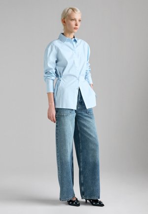 Femme aux cheveux blonds courts portant une chemise bleu clair boutonnée, un jean bleu à jambes larges et des chaussures à talons noirs avec des ornements.