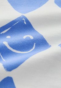 Tela de algodón con un fondo blanco y formas abstractas en azul claro, además de un patrón de caras sonrientes. Textura suave, colores vibrantes.