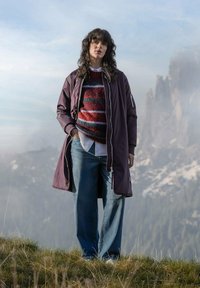 Langer lila Mantel über einem gestreiften rot-weißen Pullover, kombiniert mit weiten blauen Jeans. Stehend auf Gras mit einem Berg-Hintergrund.