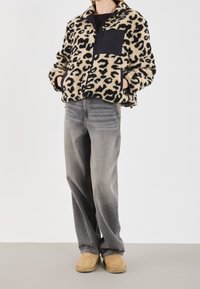 Kurtka polarowa w wzór panterki z czarną kieszenią na piersi, zestawiona z wyblakłymi szarymi dżinsami i beżowymi butami wsuwanymi, prezentująca luźny krój.