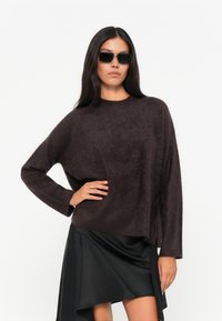 Maglione lavorato a maglia marrone scuro e peloso con scollo a girocollo e maniche lunghe, abbinato a una gonna nera asimmetrica. Occhiali da sole neri eleganti.
