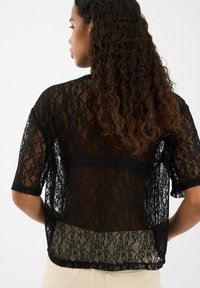 Melnā mežģīņu blūze ar īsām piedurknēm, caurspīdīgu tekstūru, sarežģītu ziedu rakstu un brīvu piegriezumu. Aizmugure ir nedaudz garāka par fronti.