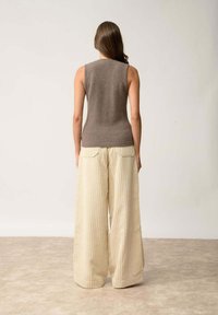 Brązowy bezrękawnik w zestawieniu z szerokimi spodniami w kolorze kremowym, w pionowe paski, z tylnymi kieszeniami, przedstawiony z tyłu.