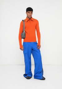 Camisola de malha laranja de mangas longas com gola de fecho, combinada com calças cargo azuis de perna larga com fechos laterais e bolsos, complementada por uma mala cinza.