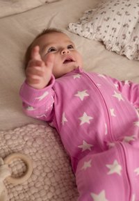 Body de bébé rose avec des motifs d'étoiles blanches et une fermeture éclair à l'avant, posé sur une couverture douce et texturée. En arrière-plan, des coussins à motifs.