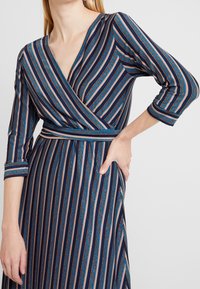 Robe portefeuille en tissu navy avec des rayures verticales roses, bleues et orange métallique. Manches longues et taille ajustée avec une ceinture.