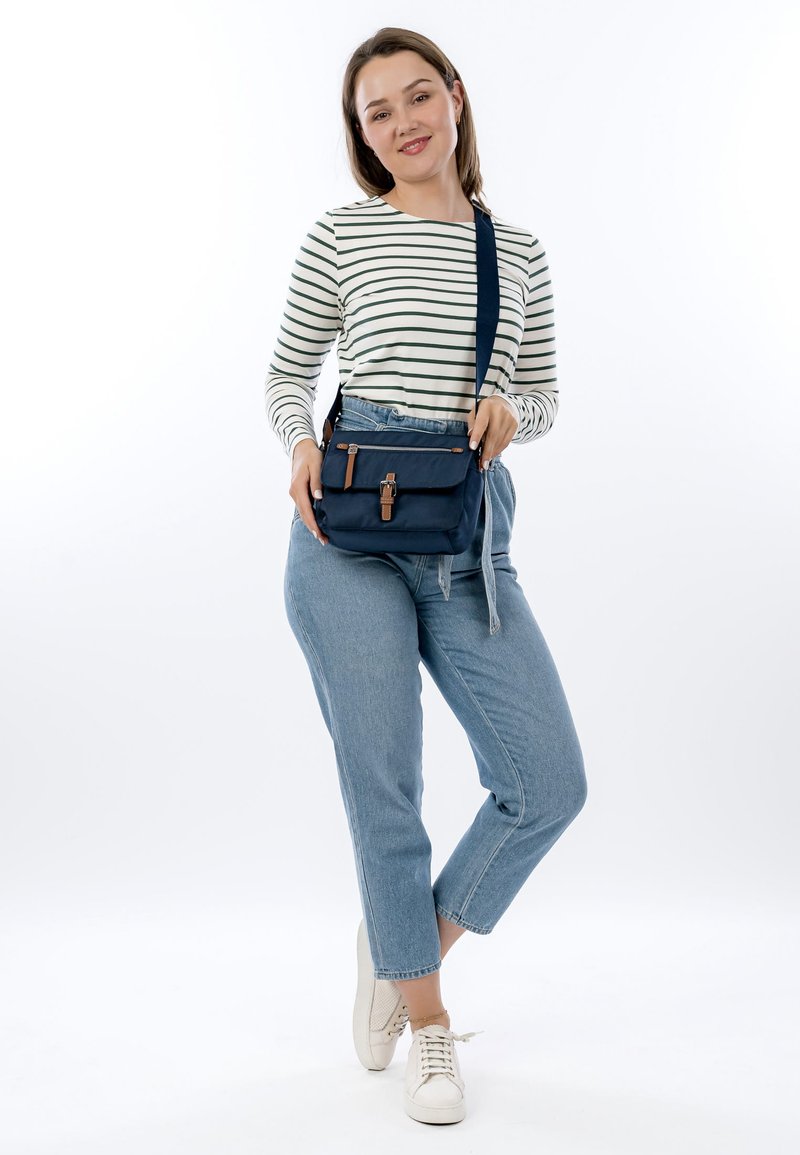 Frau steht und trägt ein gestreiftes Langarmshirt, hellblaue Jeans, weiße Turnschuhe und eine marineblaue Umhängetasche mit Lederdetails.