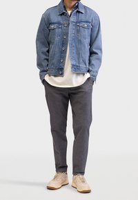 Lichte blauwe denim jacket met knopen aan de voorkant, twee borstzakken, gecombineerd met een wit T-shirt en donkergrijze geblokte broek, aangevuld met beige sneakers.