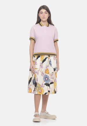 Donna in piedi che indossa una polo lilla con strisce nere e gialle senape e una gonna crema con una grande fantasia floreale colorata e scarpe platform beige.