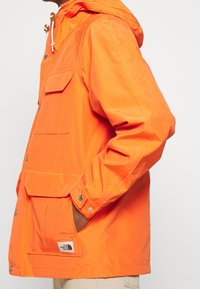 Oranžová lehká bunda s kapucí, vybavená knoflíkovými západkami, velkými předními kapsami a malou nášivkou s logem na lemu.