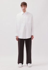 Un giovane uomo sta in piedi di fronte, indossando una camicia bianca oversize con bottoni, pantaloni neri larghi e sneakers bianche, su uno sfondo neutro.