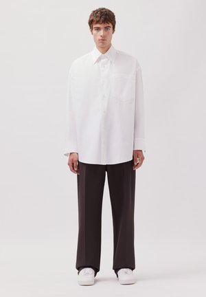 Un giovane uomo sta in piedi di fronte, indossando una camicia bianca oversize con bottoni, pantaloni neri larghi e sneakers bianche, su uno sfondo neutro.