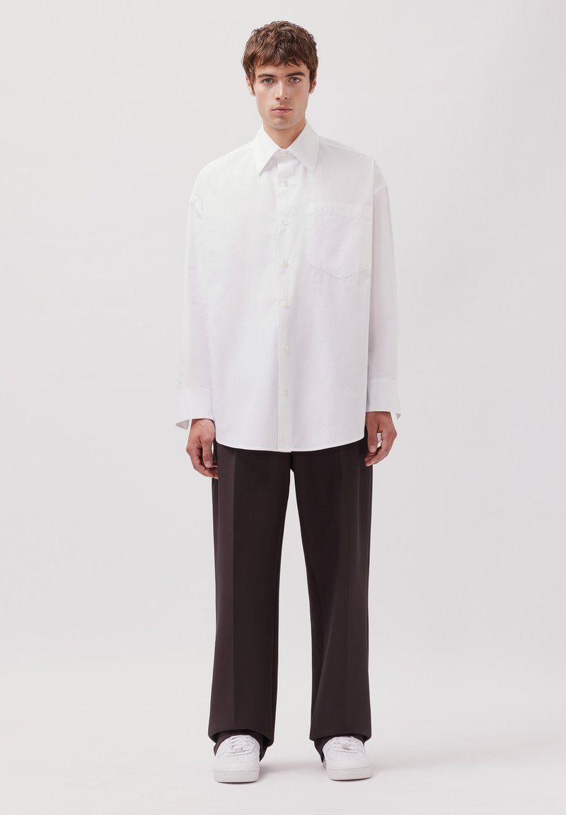 Un giovane uomo sta in piedi di fronte, indossando una camicia bianca oversize con bottoni, pantaloni neri larghi e sneakers bianche, su uno sfondo neutro.