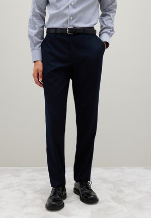 Pantaloni da cerimonia blu realizzati in tessuto con una texture liscia, abbinati a una cintura in pelle nera e scarpe neri stringate. Camicia a righe a maniche lunghe.