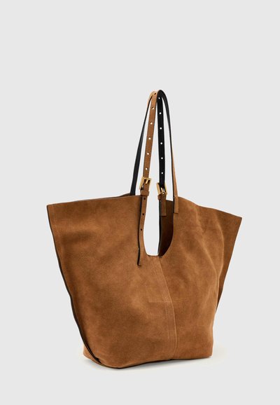 Sac fourre-tout en daim marron avec des bretelles noires réglables et des détails de boucle dorée sur un fond gris clair uni.