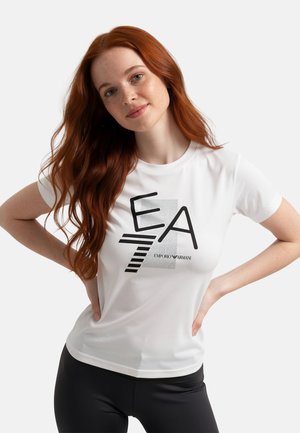 Giovane donna con lunghi capelli rossi che indossa una maglietta bianca Emporio Armani EA7 e pantaloni neri, in piedi con le mani sui fianchi.