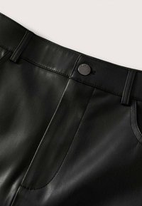 Gros plan sur un pantalon noir en cuir synthétique montrant la ceinture, le bouton, les passants de ceinture et les détails de la couture avant sur un fond clair.