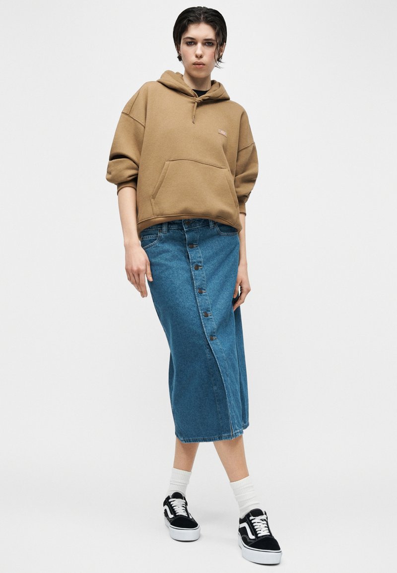 Person trägt einen braunen Hoodie, einen geknöpften blauen Jeans-Midirock, weiße Socken und schwarz-weiße Sneakers und steht vor einem einfarbigen Hintergrund.