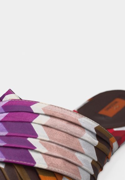 Gros plan d'une sandale à glissière multicolore avec des lanières en tissu plié aux motifs géométriques violets, roses, blancs et marron sur un fond neutre.