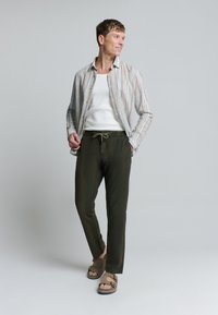 Lichtgestreepte katoenen overhemd met knopen, witte tanktop, olijfgroene broek met koord, en bruine sandalen met gespen.