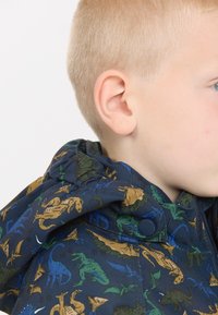 Marineblauwe jas met dinosaurussenprint in groen, bruin en blauw. Voorzien van een capuchon en drukknoopsluiting, gemaakt van gestructureerde stof.