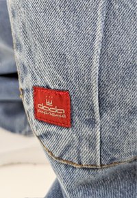 Denimstoff in Hellblau, versehen mit einem auffälligen roten Etikett, auf dem "dada supreme" in weißer Schrift und ein Krone-Logo gestickt sind.