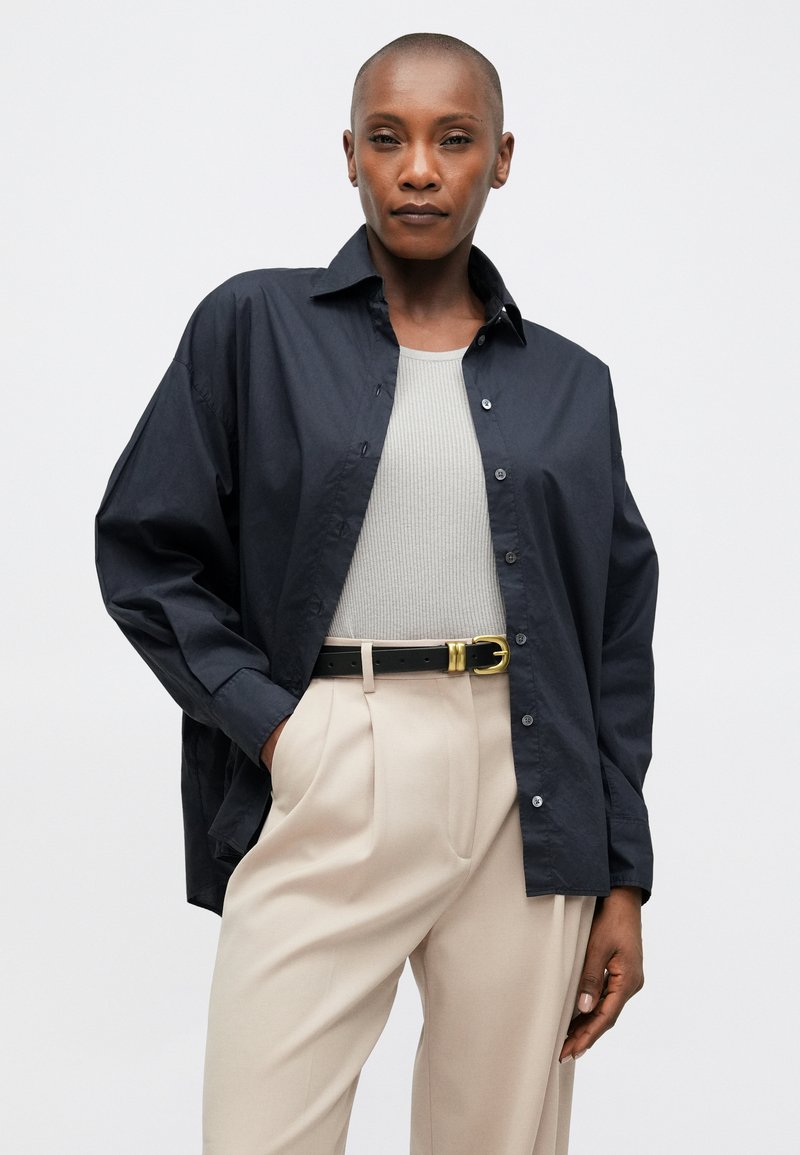 Camisa azul marino de corte relajado, con botones en el frente y mangas largas, combinada con una camiseta gris de canalé, pantalones beige y un cinturón negro con herrajes dorados.