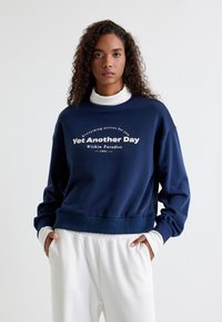 Sudadera azul marino con cuello redondo y puños acanalados. Presenta texto blanco que dice "Solo Otro Día en el Paraíso." Textura suave.