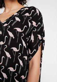 Schwarzes Oberteil aus Stoff mit einem Flamingo-Druck in Pink und Blau, V-Ausschnitt und kurzen Ärmeln. Leichtes Material mit lockerem Schnitt.