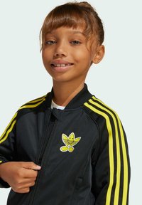 Black zip-up jacket with yellow stripes on sleeves, featuring a cartoonish logo on the chest. Smooth texture, athletic design.