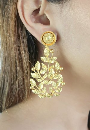 Boucle d'oreille pendante en ton or avec motif floral et feuilles, portée sur l'oreille, présentant une puce circulaire texturée et un design détaillé de feuilles et baies.