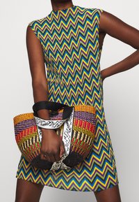 Ärmelloses Kleid mit geometrischem Muster in Blau, Grün, Gelb und Lila. Geflochtene, mehrfarbige Tasche mit schwarzem Riemen und Verzierungen.