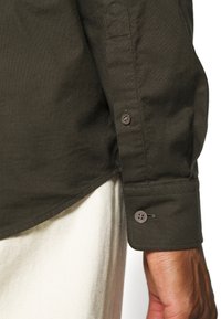 Camicia verde scuro con bottoni, presenta un polsino visibile con due bottoni, realizzata in tessuto testurizzato. Il polsino include una linguetta per la regolazione.