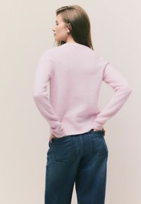 Halaszöld, hosszú ujjú kötött pulóver bordázott mandzsettával, sötétkék farmernadrággal, amely hátsó zsebekkel rendelkezik.