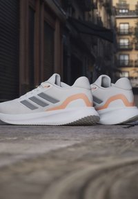 Paire de chaussures de course gris clair avec des rayures grises et des accents pêche reposant sur un pavement en pierre dans un cadre urbain.