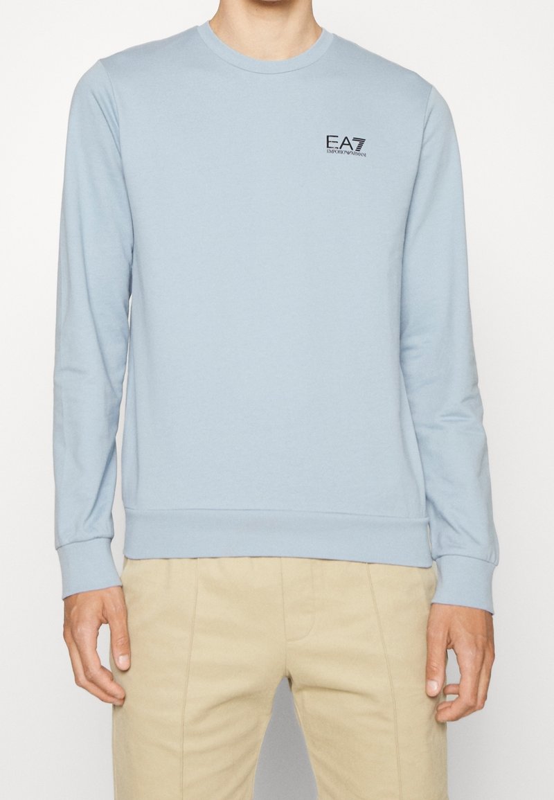 Man som bär en ljusblå långärmad sweatshirt med en liten svart EA7 Emporio Armani-logotyp på bröstet, i kombination med beige byxor.