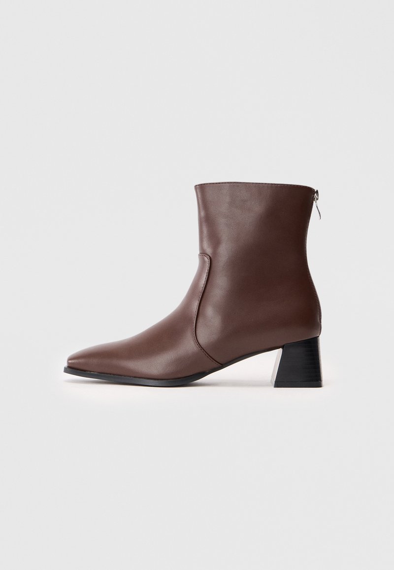 Bota de tobillo de cuero marrón con punta afilada, tacón de bloque de altura media y cremallera lateral. Textura suave y diseño minimalista.