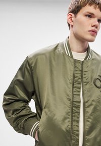 Sõjaväeroheline bomberjakk läikivas kangas esitatud esilukuga, triibulise soonikkoega kraega ja rinnale tikitud logodetailiga.