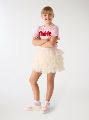 Ragazza giovane che sorride con le braccia incrociate, indossa una maglietta rosa con testo rosso, una gonna in tulle crema a strati, calzini bianchi e scarpe da ginnastica pastello.