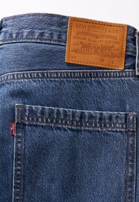 Niebieskie dżinsy z denimu z brązową skórzaną łatą z wytłoczonym tekstem, pomarańczowymi szwami i czerwoną metką Levi's na tylnej kieszeni.