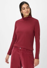 Burgunderfarbener langärmeliger Rollkragenpullover, weicher Stoff, figurbetonter Schnitt, mit subtilen Akzentdetails an den Ärmeln. Kombiniert mit passenden Cordhosen.