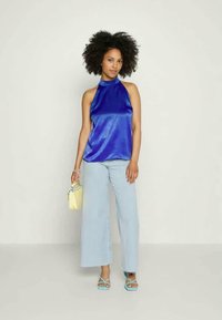 Top halter azul em cetim combinado com jeans de perna larga azul claro. Modelo segura uma pequena mala amarela, usando sandálias teal com tiras finas.