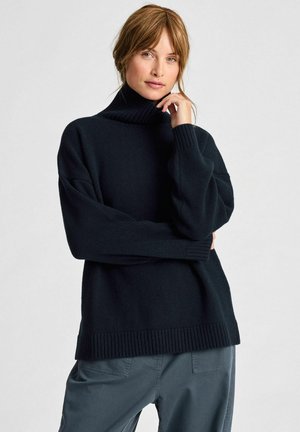 Vrouw met lichtbruin haar draagt een zwarte turtleneck trui en grijze broek, terwijl ze met één hand bij haar kin poseert tegen een effen achtergrond.