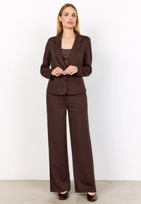 Woman standing wearing dark brown tailored blazer, matching wide-leg pants, and dark polished shoes against plain white background.