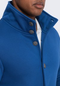 Chaqueta azul de cuello alto con una superficie texturizada, que presenta cinco botones oscuros y un forro interior blanco. Vista en primer plano centrada en el área del cuello.